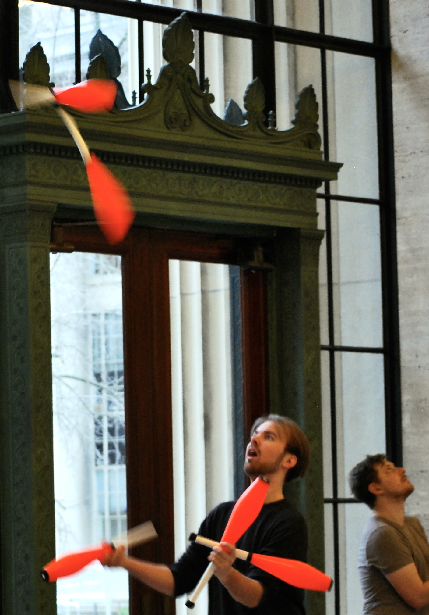 A white man with brown hair and a goatee in a black shirt and black pants juggles orange clubs: three in his hands and two in the air.