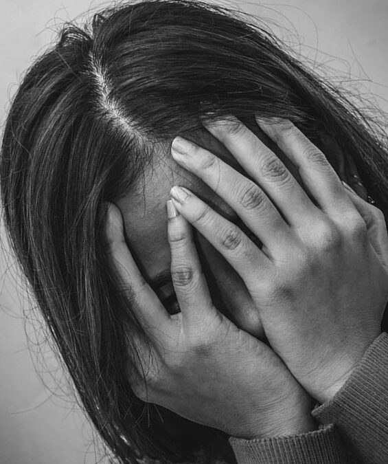 A teenager with long hair covers their face with their hands in a black-and-white photo tilted at an angle.